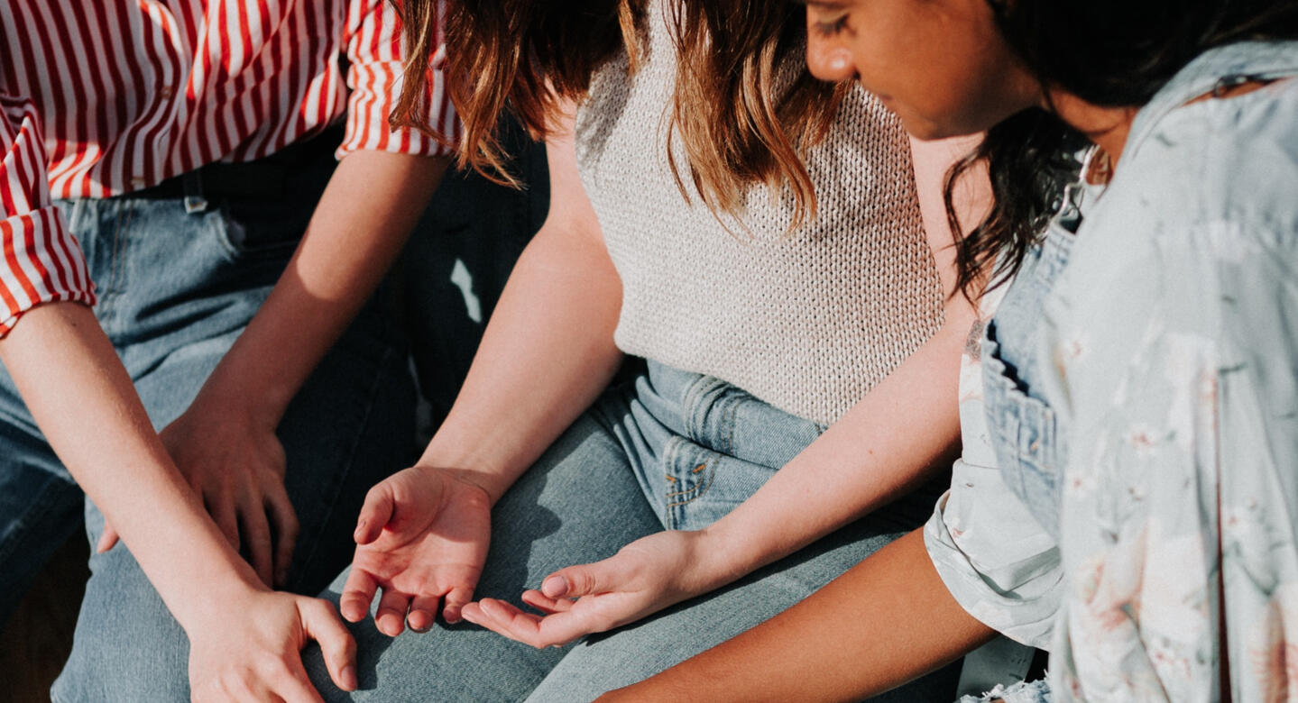 Three women sitting together. Center woman has her hands open in grief and the other two women comfort her.