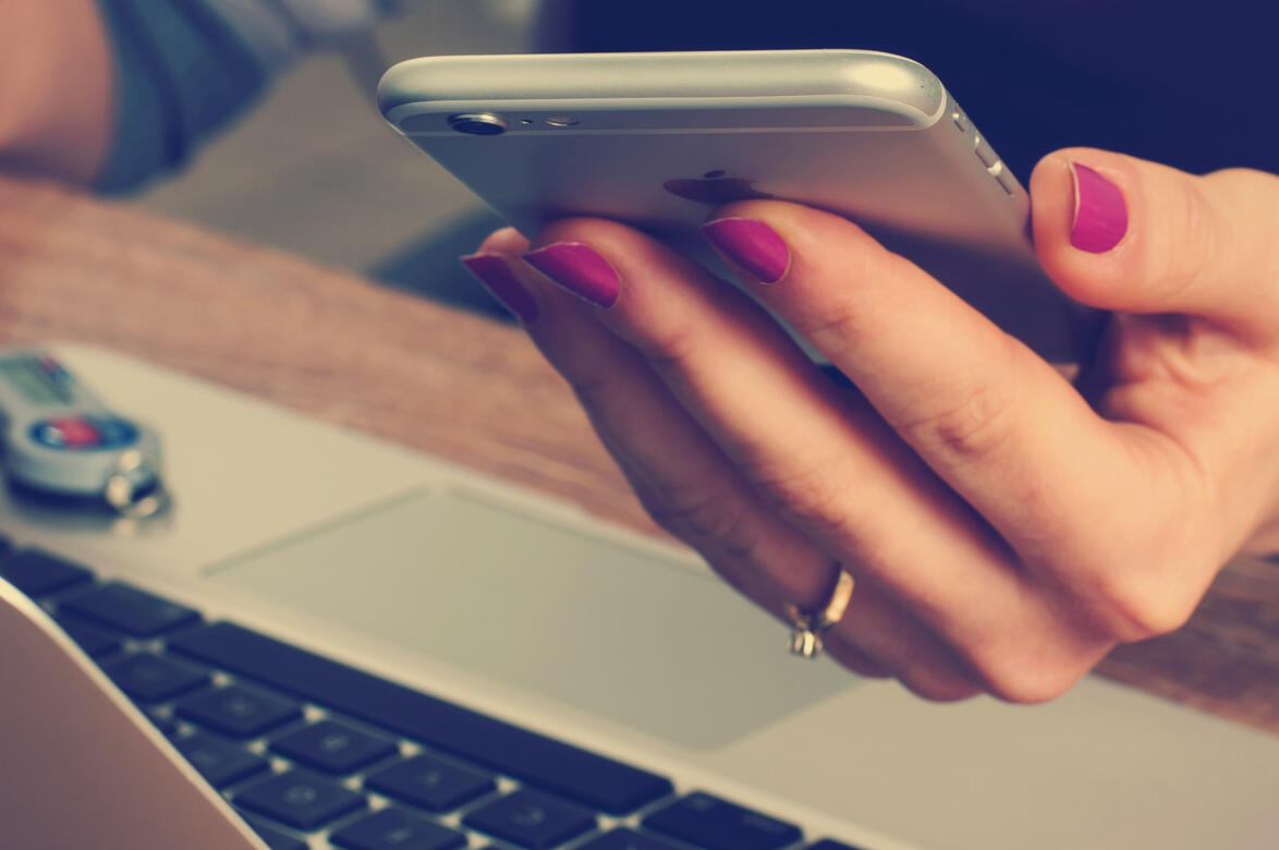 Untitled A woman with pink fingernails holding a cell phone in her left hand while sitting in front of a laptop.
